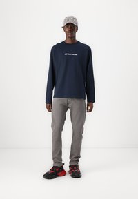 Camisa de manga larga azul marino con "RED BULL RACING" impreso en blanco, jeans grises, y zapatillas negras con acentos rojos, combinadas con una gorra gris claro.