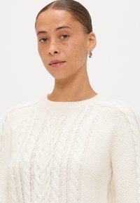 Pullover a collo rotondo in maglia cable-knit color crema, caratterizzato da motivi a maglia testurizzati. Tessuto morbido e liscio con una vestibilità comoda.