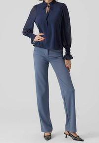 Blouse bleu marine à manches longues avec un décolleté en goutte d'eau, associée à un pantalon bleu clair. Des talons à motifs noirs et blancs complètent la tenue.