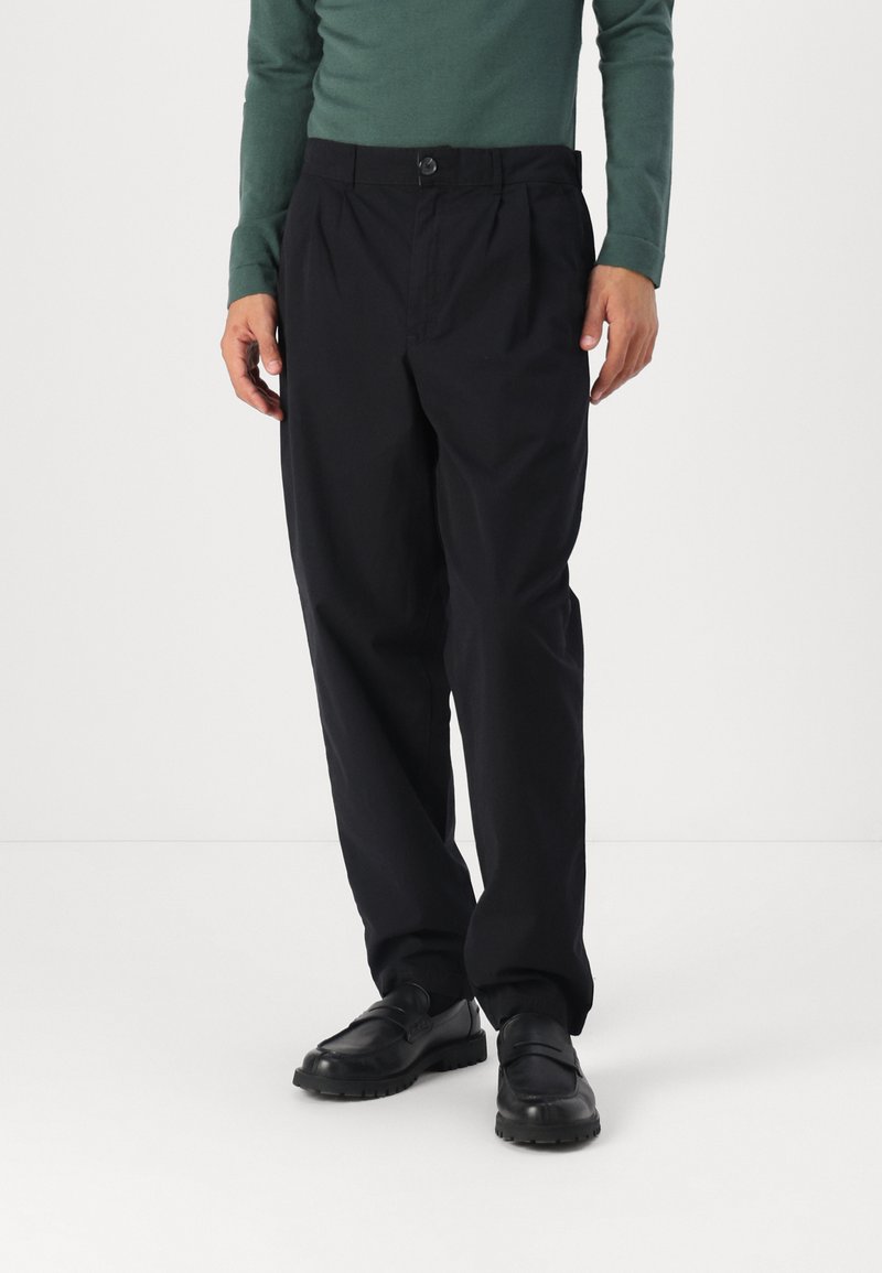 Zwarte geplooide broeken met een knoopsluiting, voorzien van zijzakken en een relaxte pasvorm. G gedragen met zwarte schoenen en een groene top.