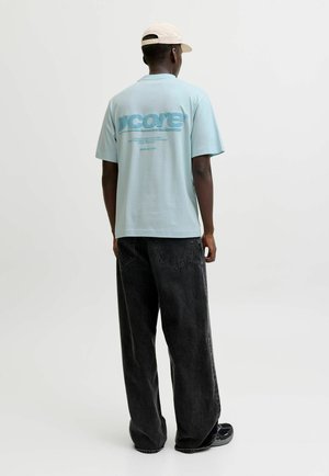 Ung mand iført en lyseblå t-shirt med teksten "ycore" på ryggen, sorte vidde jeans, sorte sneakers og en beige kasket, set bagfra.