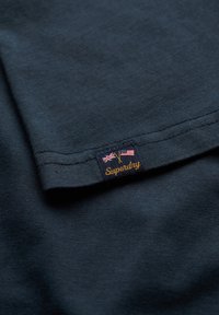 Mørk marineblå bomulds T-shirt med en glat tekstur, der har et broderet mærke med UK- og US-flag samt "Superdry" i orange skrift.