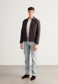 Tamnosiva teksturirana jakna s ovratnikom i prednjim džepom, nošena preko bijele košulje; svijetloplave traperice i crne tenisice upotpunjuju odjevnu kombinaciju.