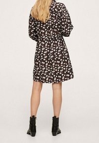 Žena s dlouhými blond vlasy, oblečená v černých květinových šatech a černých kotníkových botách, stojící zády k fotoaparátu.