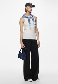 Woman wearing black cap, white sleeveless top, blue and white striped sweater draped over shoulders, black wide-leg pants, white sneakers, holding blue handbag.