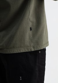 Olivgrünes Baumwoll-T-Shirt mit einem gestickten Saum und einem kleinen schwarzen Etikett mit einem weißen "J". Getragen mit schwarzen Hosen, die weiße "J"-Stiche zeigen.