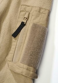 Beige fliisist dressipluus, millel on lukuga tasku ja Velcro-patch, omades tekstureeritud kangast ja tugevdusõmblusi hemmi ja mansettide lähedal.