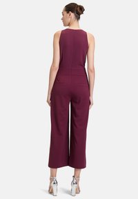 Combinaison bordeaux avec un design sans manches, corsage ajusté, silhouette à jambes larges et fermeture éclair cachée au dos. Comprend deux poches arrière.