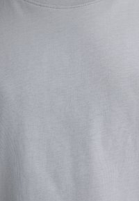 Close-up of light gray ribbed fabric on a plain crew neck garment, showing fine knit texture and subtle shadows.