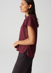 Maglietta strutturata bordeaux con maniche corte, caratterizzata da un orlo curvo e spacchi laterali, abbinata a pantaloni neri. Tessuto morbido con un motivo sottile.