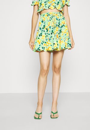 Femme portant une jupe fleurie verte et un crop top assorti, associée à des sandales vertes à talons, debout devant un fond uni.