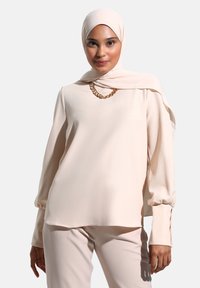 Hladký long-sleeve top v barvě blush, s vysokým výstřihem, volným střihem a detailem řetízku. V kombinaci se sladěnými kalhotami.