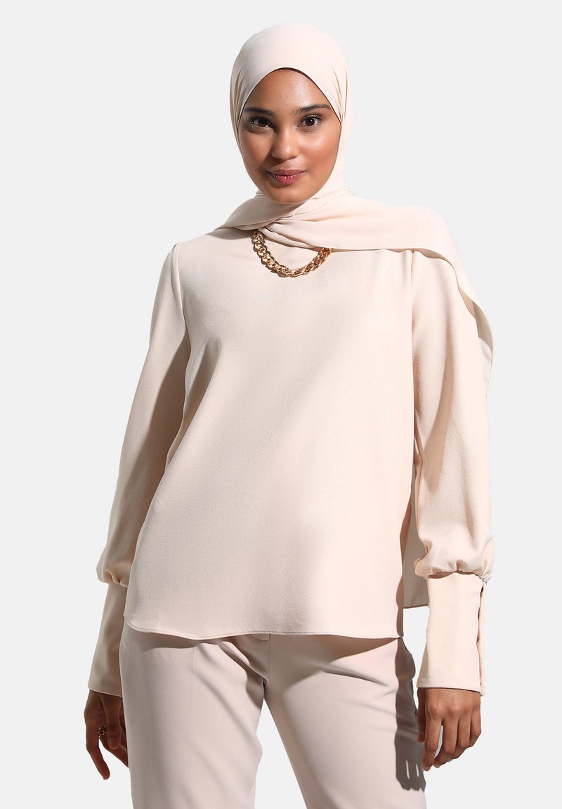 Hladký long-sleeve top v barvě blush, s vysokým výstřihem, volným střihem a detailem řetízku. V kombinaci se sladěnými kalhotami.