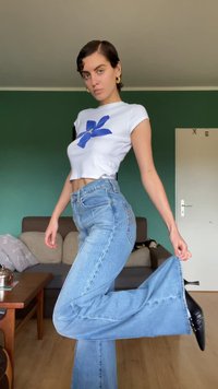 Fehér crop top kék virágmintával, magas derekú világoskék trapéznadrág és fekete magassarkú cipők, zöld fal háttér előtt.