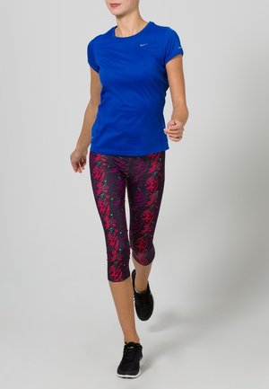 Kobieta w niebieskiej koszulce z krótkim rękawem i wzorzystych legginsach capri idzie, mając na sobie czarne buty sportowe na jednolitym jasnym tle.