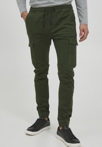 Groene cargo broek met een tapered fit, voorzien van twee zijzakken, ribgebreide boorden en een trekkoord in de taille, gecombineerd met zwarte sneakers.