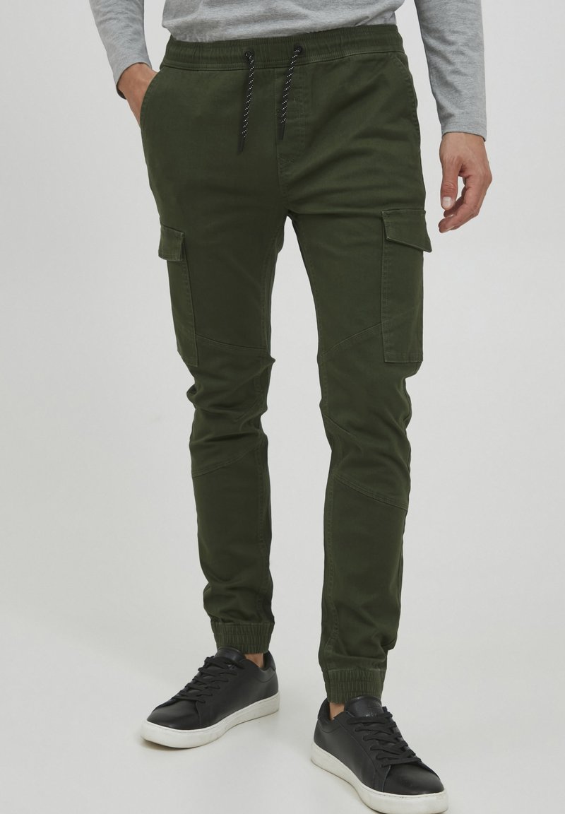 Groene cargo broek met een tapered fit, voorzien van twee zijzakken, ribgebreide boorden en een trekkoord in de taille, gecombineerd met zwarte sneakers.