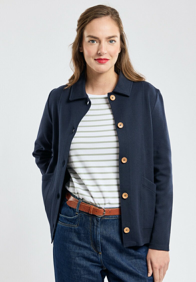 Veste bleu marine en tissu texturé, avec un col, des boutons en bois et deux poches latérales. Portée sur une chemise rayée.