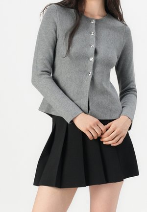 Femme portant un cardigan gris à manches longues boutonné et une jupe noire plissée, debout, les mains jointes devant elle.
