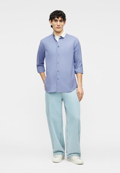 Νεαρός άνδρας στέκεται με τα χέρια στις τσέπες, φορώντας ανοιχτό μπλε πουκάμισο με ρίγες και κουμπιά, ανοιχτό μπλε τζιν με φαρδύ πόδι και λευκά αθλητικά παπούτσια.