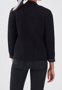 Czarna pikowana kurtka z fakturowanym wzorem, z długimi rękawami i srebrnymi akcentami na mankietach. Noszona z czarnymi spodniami.