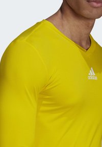 Geel sportshirt met een V-halsontwerp, gemaakt van ademend materiaal, met een diagonale geribbelde textuur en een wit Adidas-logo op de borst.