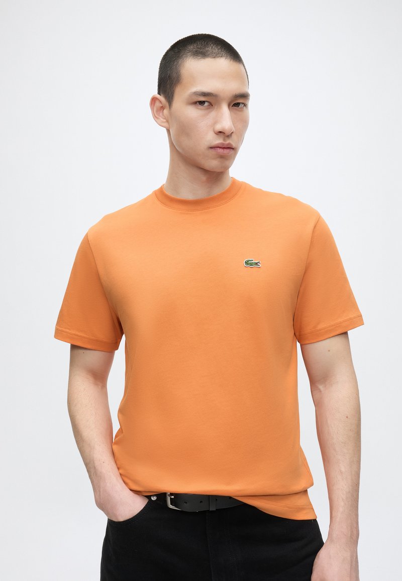 Man met kort haar die een oranje shirt met korte mouwen draagt met een klein groen krokodillenlogo op de borst, poserend met één hand in de zak.