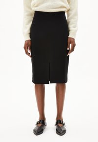 Zwarte kokerrok met een split aan de voorkant; gladde textuur, hoge taille ontwerp. Gekoppeld met zwarte schoenen met een rond metalen accent.