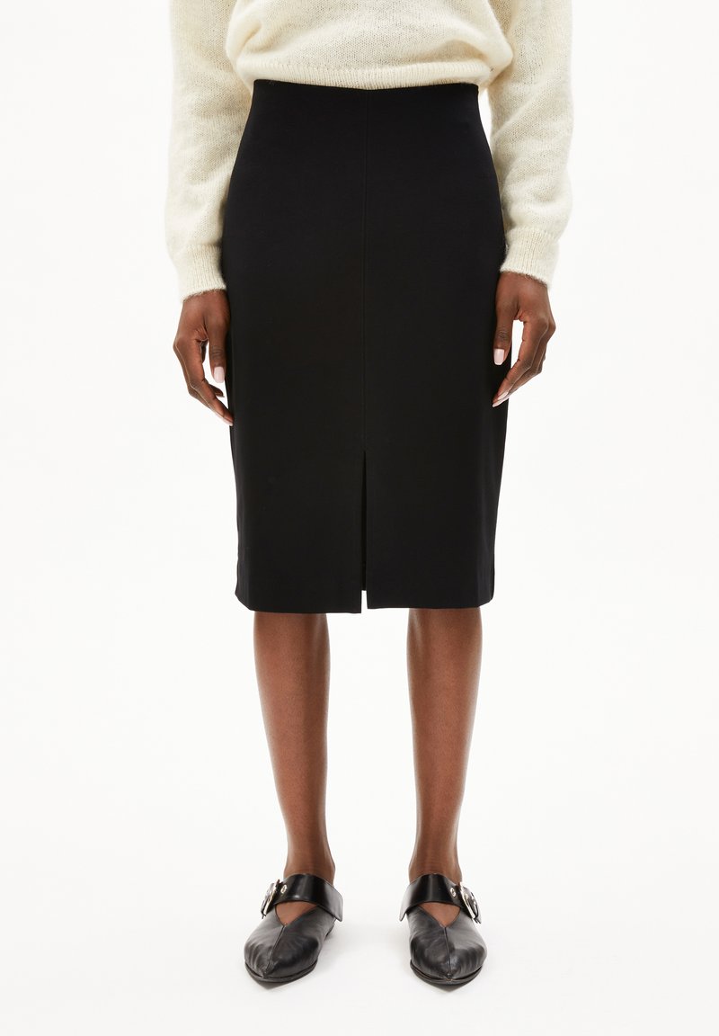 Jupe crayon noire avec une fente avant ; texture lisse, design taille haute. Associée à des chaussures noires avec un accent métallique rond.