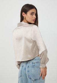 Camicia cropped in satin color beige chiaro con colletto e maniche lunghe, dotata di polsini con bottone. Indossata con jeans in denim blu a vita alta.