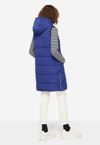 Gilet rembourré bleu avec capuche, design matelassé vertical, fentes latérales, porté sur une chemise à rayures à manches longues, associé à un pantalon blanc et des baskets.