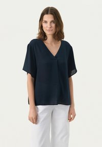 Blusa a maniche corte blu navy con scollatura a V e vestibilità ampia, realizzata in tessuto leggero. Abbinata a pantaloni bianchi a gamba larga.