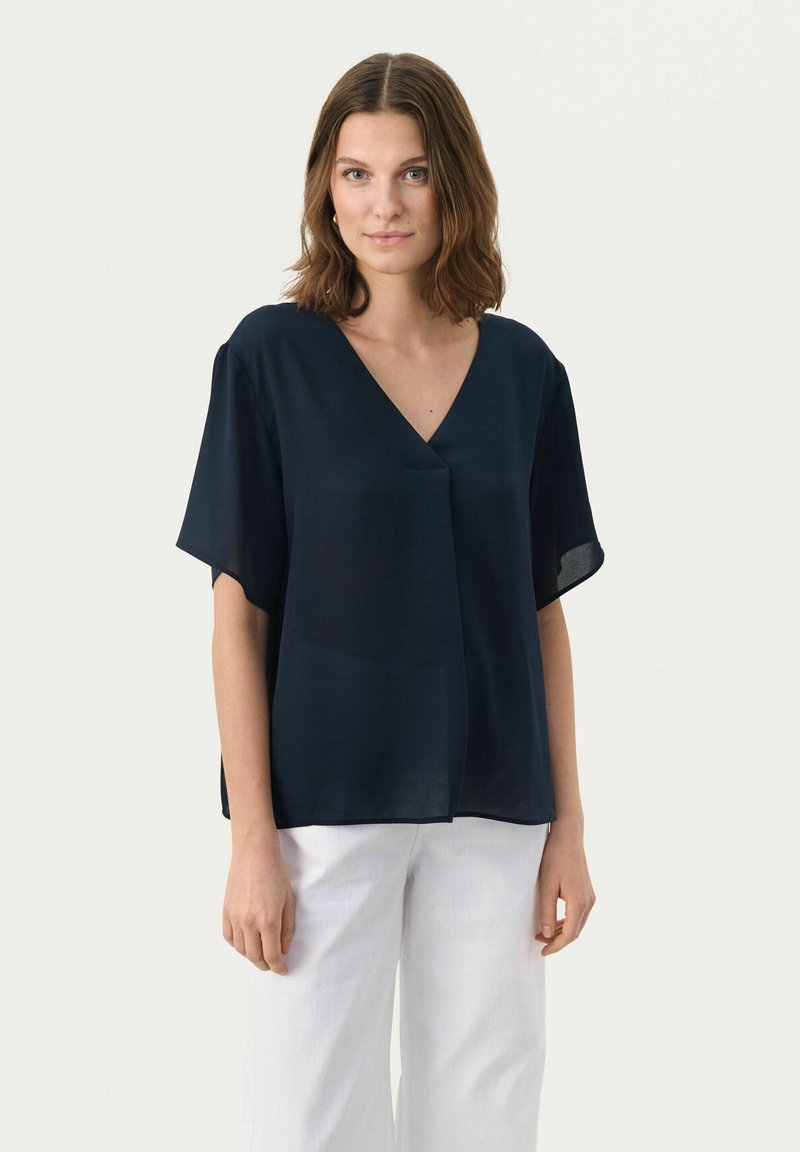 Marineblaues, kurzärmliges Blusenshirt mit V-Ausschnitt und lässiger Passform, aus leichtem Stoff gefertigt. Kombiniert mit weißen, weit geschnittenen Hosen.