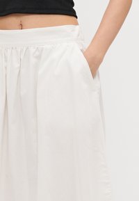 Jupe plissée blanche avec une taille élastique et des poches latérales, fabriquée en tissu lisse. Présente un design épuré et minimaliste sans motifs.