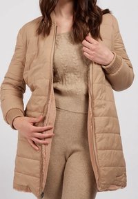 Kvinde iført beige strikket sweater- og buksesæt med en beige quiltet frakke foret med blødt stof, delvist åbnet med lynlås, hænderne justerer foran.