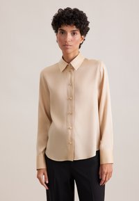 Blusa beige realizzata in tessuto morbido, con colletto classico, maniche lunghe e chiusura a bottoni con bottoni abbinati.