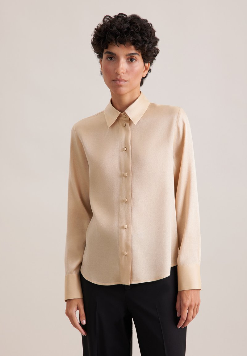 Blusa beige realizzata in tessuto morbido, con colletto classico, maniche lunghe e chiusura a bottoni con bottoni abbinati.
