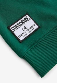 Groen sweatshirtstof met een zwartomrand etiket in het wit met de tekst: "SUB5CRIBE LA. 33.9871° N, 118.4755° W. Los Angeles, Californië."