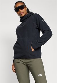 Femme portant une polaire à capuche noire, un pantalon vert olive avec le logo The North Face, et de grandes lunettes de soleil noires sur un fond uni.