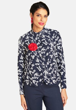 Woman wearing navy floral jacket with white blossoms, a red fabric flower brooch, matching red earrings, and dark navy pants.