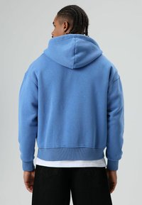 Blauwe hoodie met een losse pasvorm. Voorzien van een capuchon met trekkoord, geribbelde manchetten en zoom. Gladde stoftextuur, gedragen over een wit shirt.