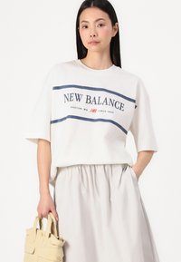 Balts kokvilnas T-krekls ar tumši zilām "NEW BALANCE" uzrakstiem un dekoratīvām strīpām, apvienots ar gaiši pelēkiem svārkiem un bēšu somu.