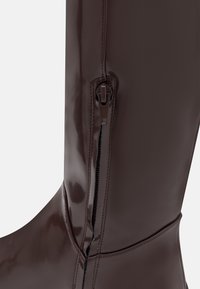 Bota marrón brillante hasta la rodilla con cremallera lateral, textura suave y un diseño ajustado. Los detalles cosidos realzan la apariencia elegante.