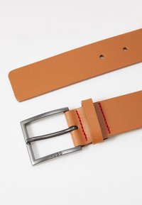 Curea din piele de culoare maro deschis, cu o cataramă rectangulară argintie, detalii cu cusături roșii și mai multe găuri de ajustare lângă capătul cataramei.