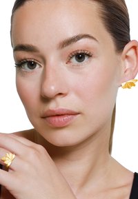 Les bijoux en ton doré présentent un design floral ; les boucles d'oreilles ont une forme de pétale, tandis que la bague reflète ce motif complexe.