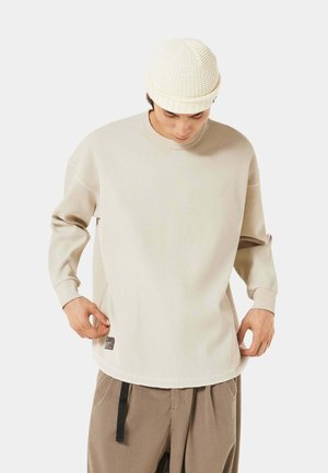 Cremfarbnes Langarm-Rippshirt mit rundem Halsausschnitt und seitlichen Schlitzen, kombiniert mit einer strukturierten cremfarbenen Mütze.