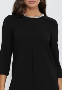 Schwarzes Kleid mit Dreiviertelärmeln, dekorativem Kragen mit silberner Beadings und einer boxy Silhouette mit einem subtilen Vordernahtdetail.