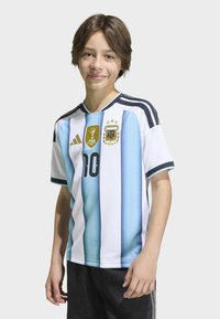 Jeune personne portant un maillot de football de l'Argentine avec le numéro 10, se tenant devant un fond uni, souriant légèrement.