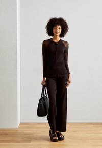 Combinaison noire avec des manches en mesh longues, fermeture éclair à l'avant, corps ajusté, jambes larges. Le mannequin tient un sac à main noir et porte des sandales à plateforme.