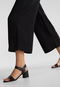 Jambes portant un pantalon large noir raccourci et des sandales noires à bout ouvert avec des talons blocs et des lanières à la cheville marchant sur un sol clair.